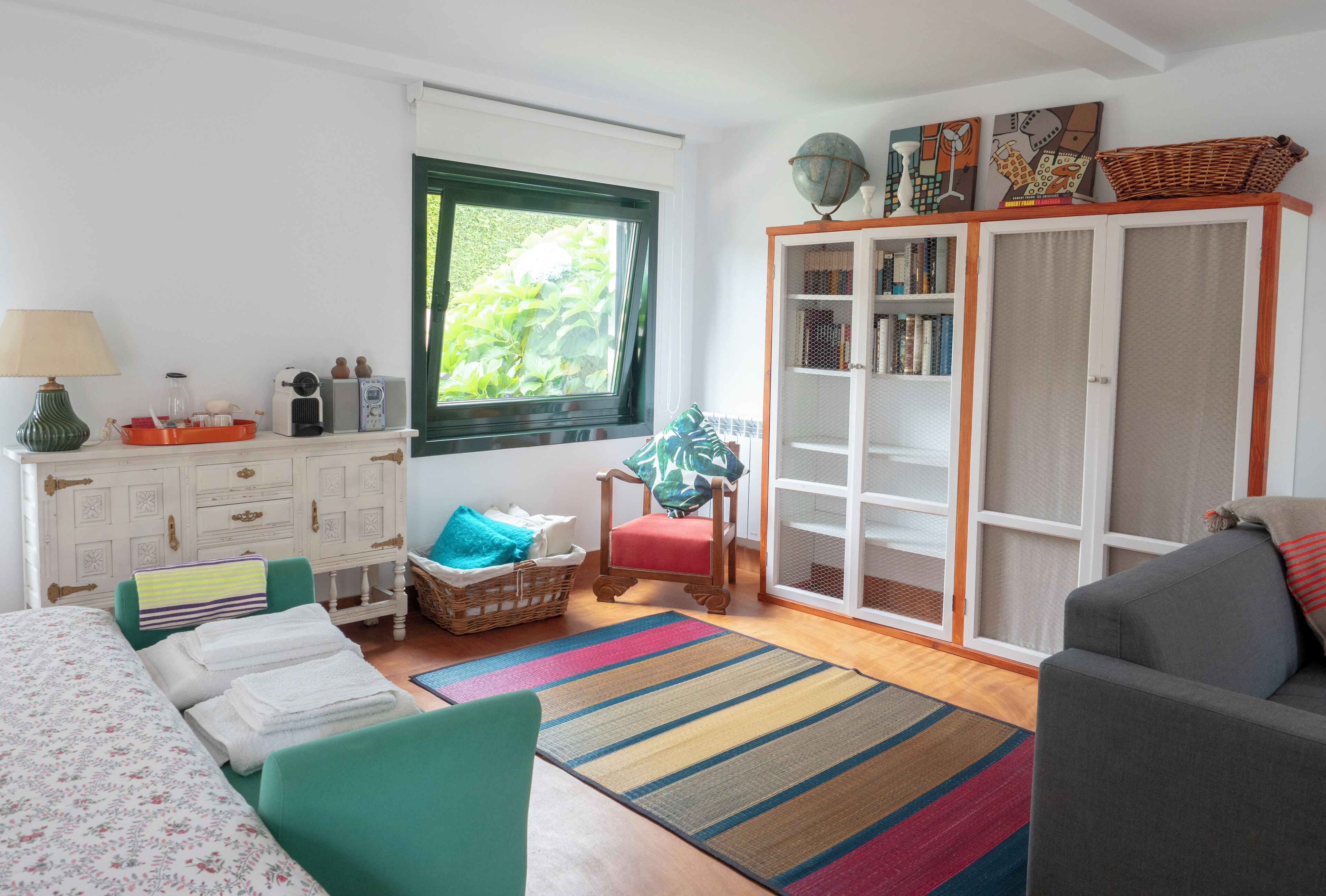 A room with a bed, a sofa, a chair, a bookshelf, and a window with a view of greenery.