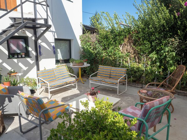 Eine Terrasse mit Gartenmöbeln, darunter zwei gestreifte Sofas und ein Korbsessel, umgeben von Topfpflanzen und einer weißen Wand.