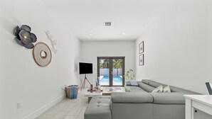 A modern living room with a sectional sofa, a flat-screen TV, and a large window overlooking a swimming pool. - Family Fun Oasis - Pool - Games - Fenced Yard &BBQ (West Park)