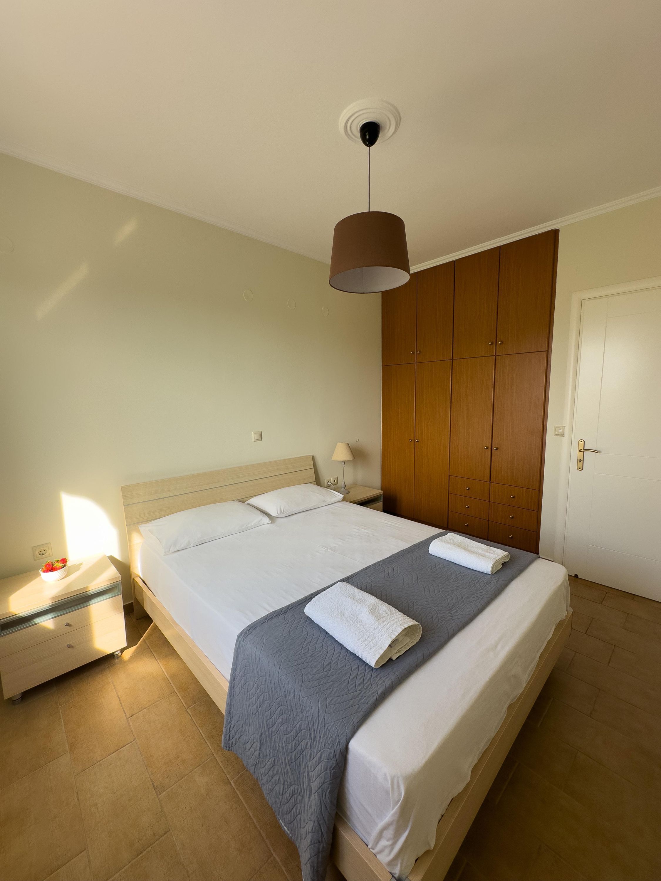 A neatly made bed with a grey blanket and two white towels, a wooden wardrobe, a bedside table with a lamp, and a ceiling light fixture.