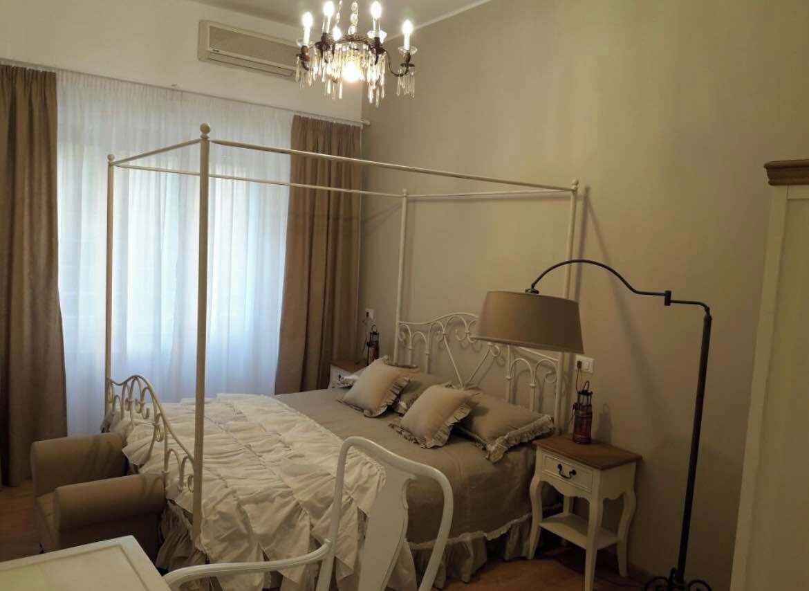 A four-poster bed with a canopy, a bedside table, a lamp, and a chair in a room with a chandelier.