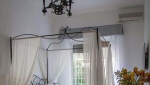 Ein Schlafzimmer mit Himmelbett, einer Chandelierleuchtre, einem Nachttisch, einem Sessel und einer Vase mit Blumen.