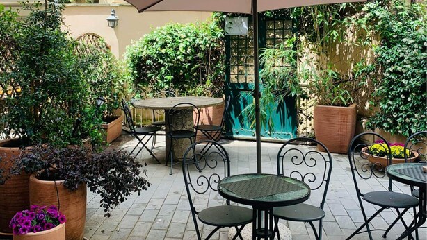 Eine Terrasse im Freien mit Tischen und Stühlen, umgeben von Blumentöpfen und einem großen Sonnenschirm.