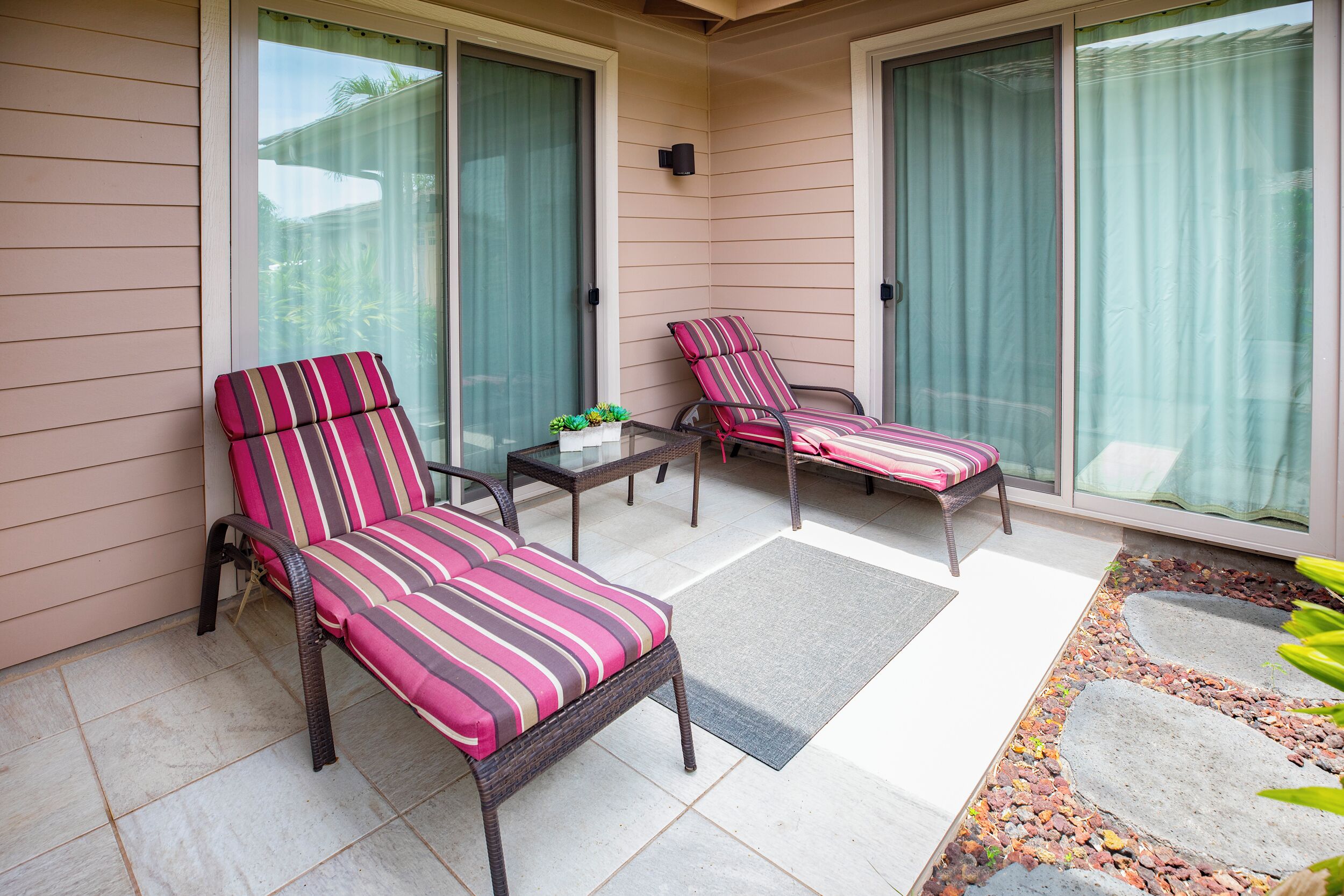 A covered patio area with two striped lounge chairs, a small table with a plant, and a decorative stone border.