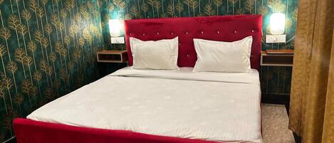 A bed with a red headboard and white bedding in a room with patterned wallpaper and a nightstand with a lamp.