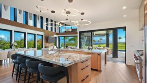 A modern kitchen with a large island, bar stools, and a view of the outdoors. - Luxury 6BR Hale o ka Nene Estate w/ Pool/Spa & Golf Views, Near Beach (Koloa)