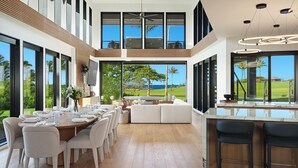 A modern kitchen with a large island, a dining area with a wooden table and chairs, and large windows offering a view of the outdoors. - Luxury 6BR Hale o ka Nene Estate w/ Pool/Spa & Golf Views, Near Beach (Koloa)