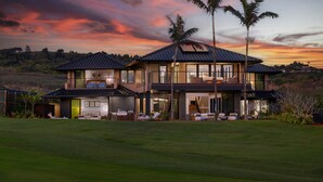 A modern two-story house with a large front lawn, a covered outdoor seating area, and a view of the sunset. - Luxury 6BR Hale o ka Nene Estate w/ Pool/Spa & Golf Views, Near Beach (Koloa)