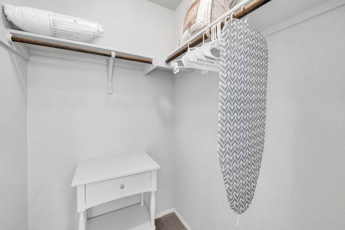 A white closet with a wooden shelf, a white bedside table, and a patterned shower curtain.