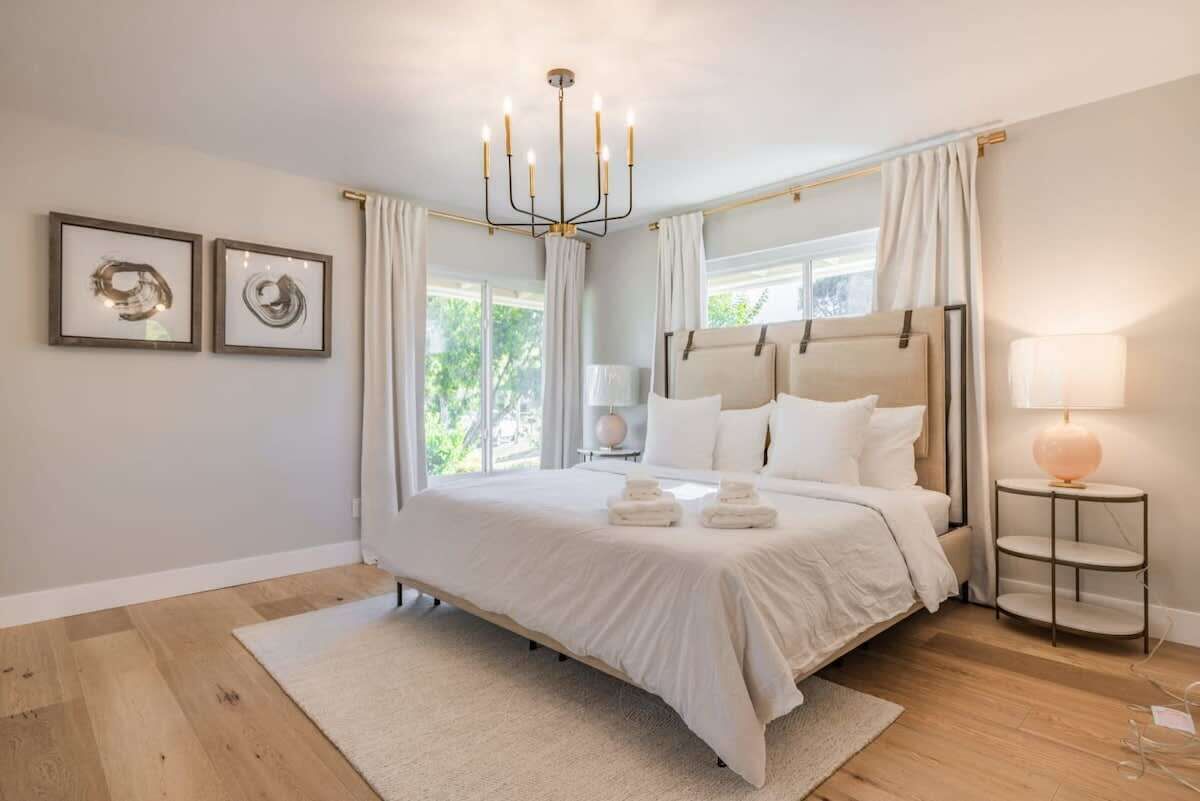 A spacious bedroom with a large bed, a chandelier, a bedside table with a lamp, and two framed artworks on the wall.