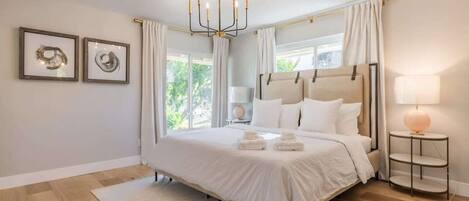 A spacious bedroom with a large bed, a chandelier, a bedside table with a lamp, and two framed artworks on the wall.