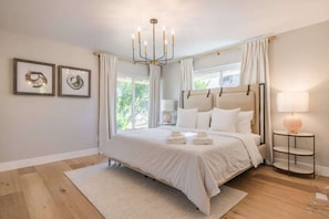 A spacious bedroom with a large bed, a chandelier, a bedside table with a lamp, and two framed artworks on the wall. - @ Marbella Lane - Cozy Modern Home w/ Relaxing Design & Pool (Fullerton)