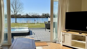 Ein Wohnzimmer mit großem Fernseher, einem Holztisch und Blick auf einen See und eine Außenwhirlpool.