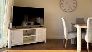 Ein Wohnzimmer mit Flachbildfernseher auf einer Holzkommode, einer runden Wanduhr und einem Esstisch mit zwei Stühlen.