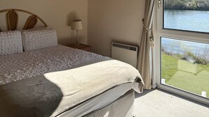 A bedroom with a wooden ceiling, a bed with a grey blanket, a bedside lamp, and a window with a view of a lake.