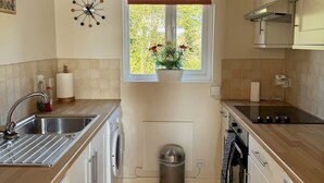 A compact kitchen with a washing machine, a trash bin, and a window with a view of greenery.