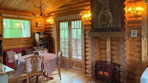 A cozy log cabin interior with a dining area, a fireplace with a deer head above it, and a large window overlooking greenery.