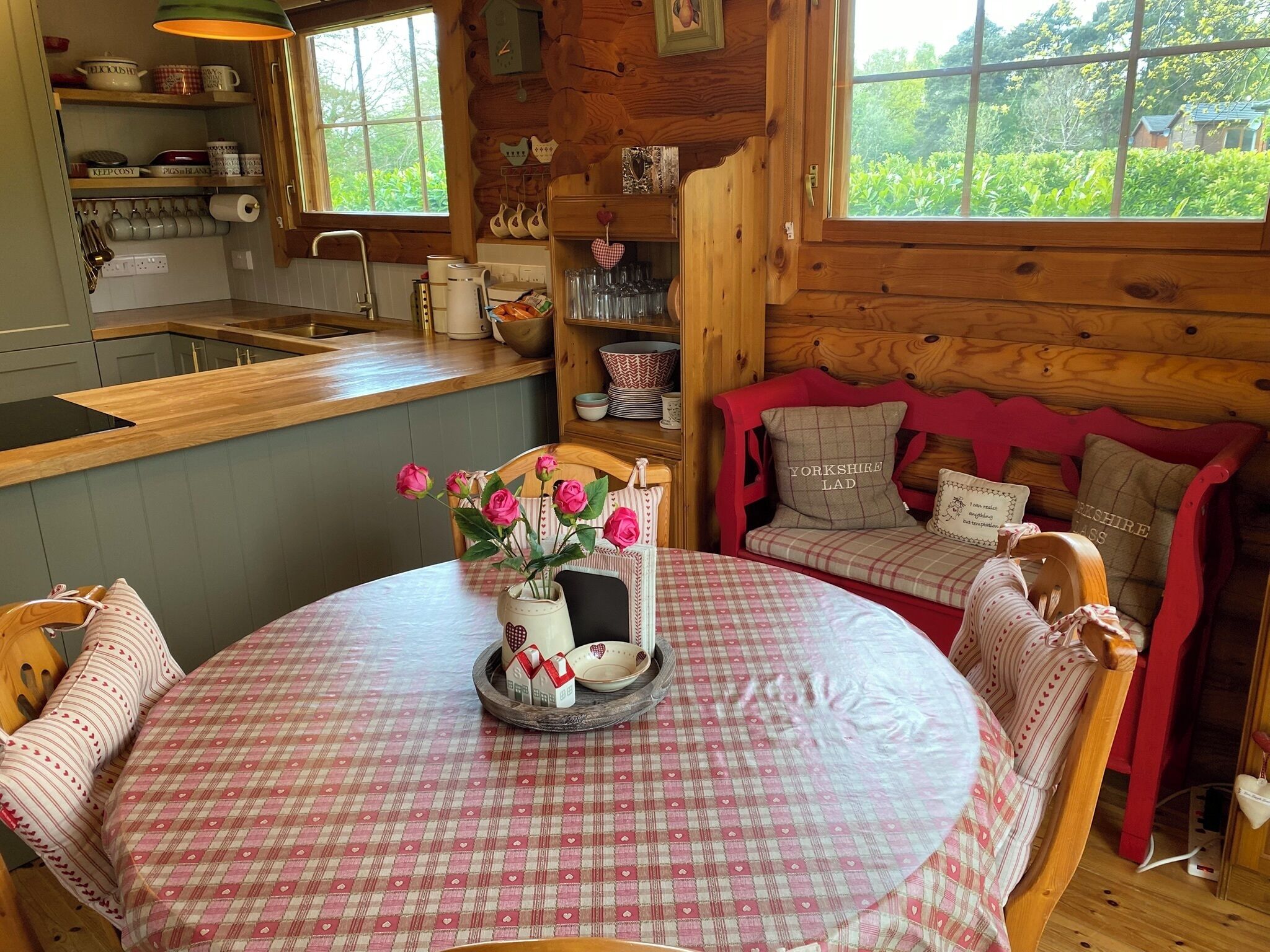 A cozy cabin interior with a wooden dining table, a red sofa, and a window overlooking greenery.