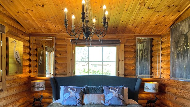 A log cabin bedroom with a large bed, wooden walls, a chandelier, and framed wildlife paintings.