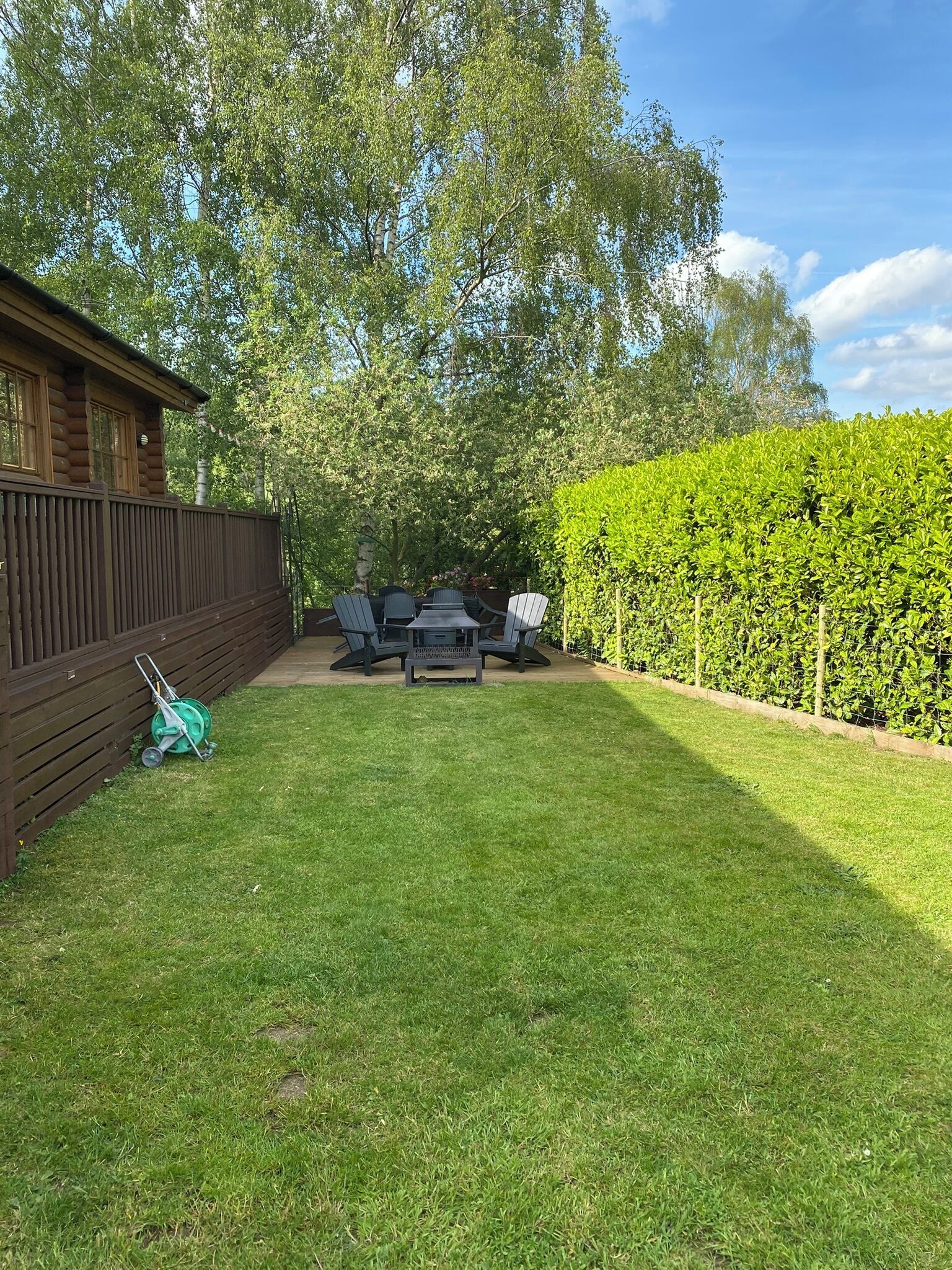 Ein Garten mit Holzterrasse, Rasenfläche, Hecke und Gartenmöbeln.