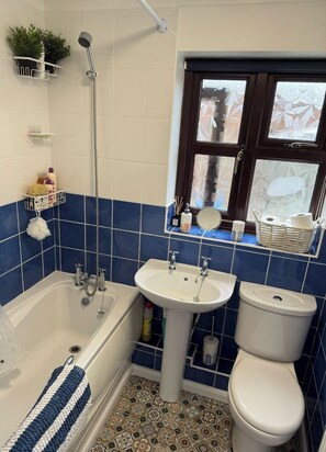 A bathroom with a bathtub, sink, and toilet, blue and white tiles, a window with frosted glass, and a shelf with toiletries.