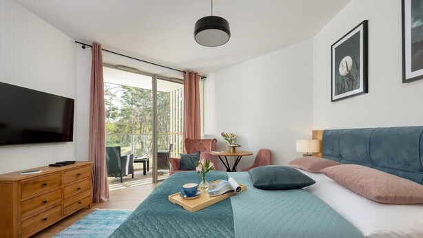 A modern bedroom with a bed, a TV, a desk, and a view of the outdoors.