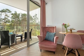 Una habitación moderna con un gran ventanal corredero que da a un espacio exterior, un sillón rosa, una mesa redonda de madera con un jarrón de flores y una mesa de comedor con sillas.