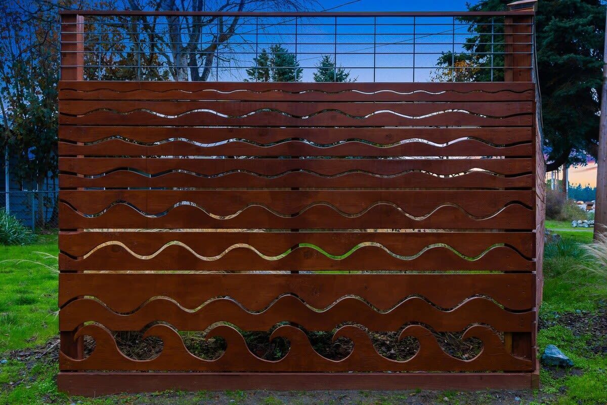 A wooden structure with wavy cutouts, possibly a decorative panel or fence, set in a grassy area with trees and a clear sky in the background.