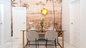 Un coin repas avec une table en verre, deux chaises et une fresque murale représentant des palmiers.
