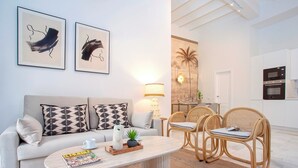 Un salon moderne avec un canapé, une table basse et des fauteuils en rotin. On peut voir un magazine sur la table, une lampe et une œuvre d’art au mur.