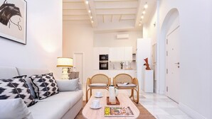Un salon moderne avec un canapé, une table basse et deux fauteuils. On y trouve une table à manger avec un magazine et un vase. Les murs de la pièce sont blancs et le plafond est doté d’un éclairage encastré.