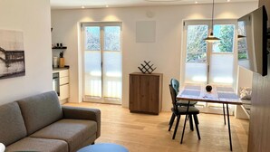 Ein modernes Wohnzimmer mit einer Couch, einem Esstisch und Stühlen. Im Hintergrund steht ein Holzschrank und eine Küchenzeile.