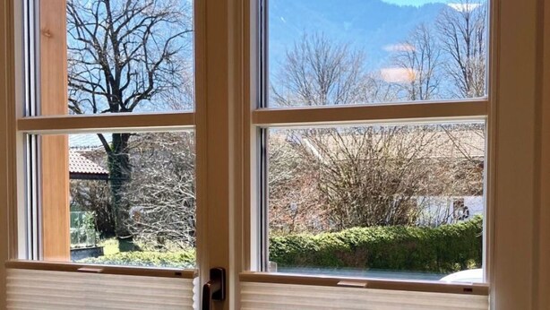 Ein Zimmer mit großem Fenster, einem Holztisch und Blick auf Berge und Bäume.