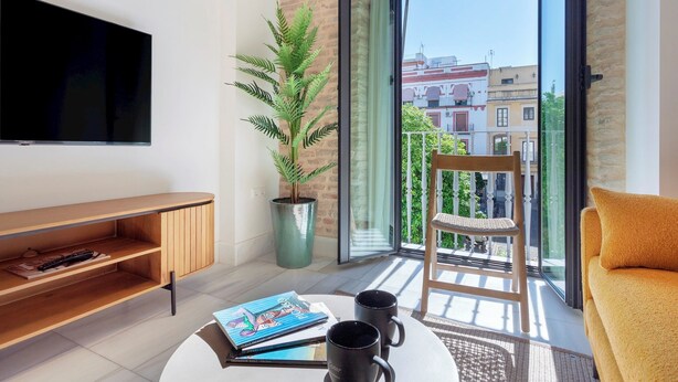 A modern living room with a flat-screen TV, a wooden entertainment unit, a potted plant, a round coffee table with books and mugs, a yellow sofa, and a balcony with a view of buildings. - Genteel Home San Vicente III (Seville)