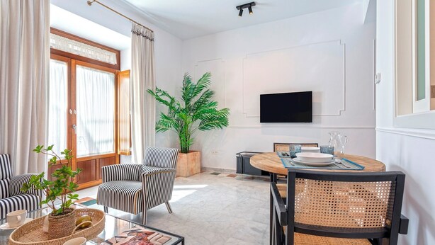 A modern living room with a dining area, featuring striped chairs, a large potted plant, and a flat-screen TV on the wall.