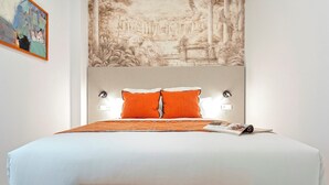 A neatly made bed with white bedding and two orange pillows, a wooden nightstand, and a wall tapestry depicting a classical scene.
