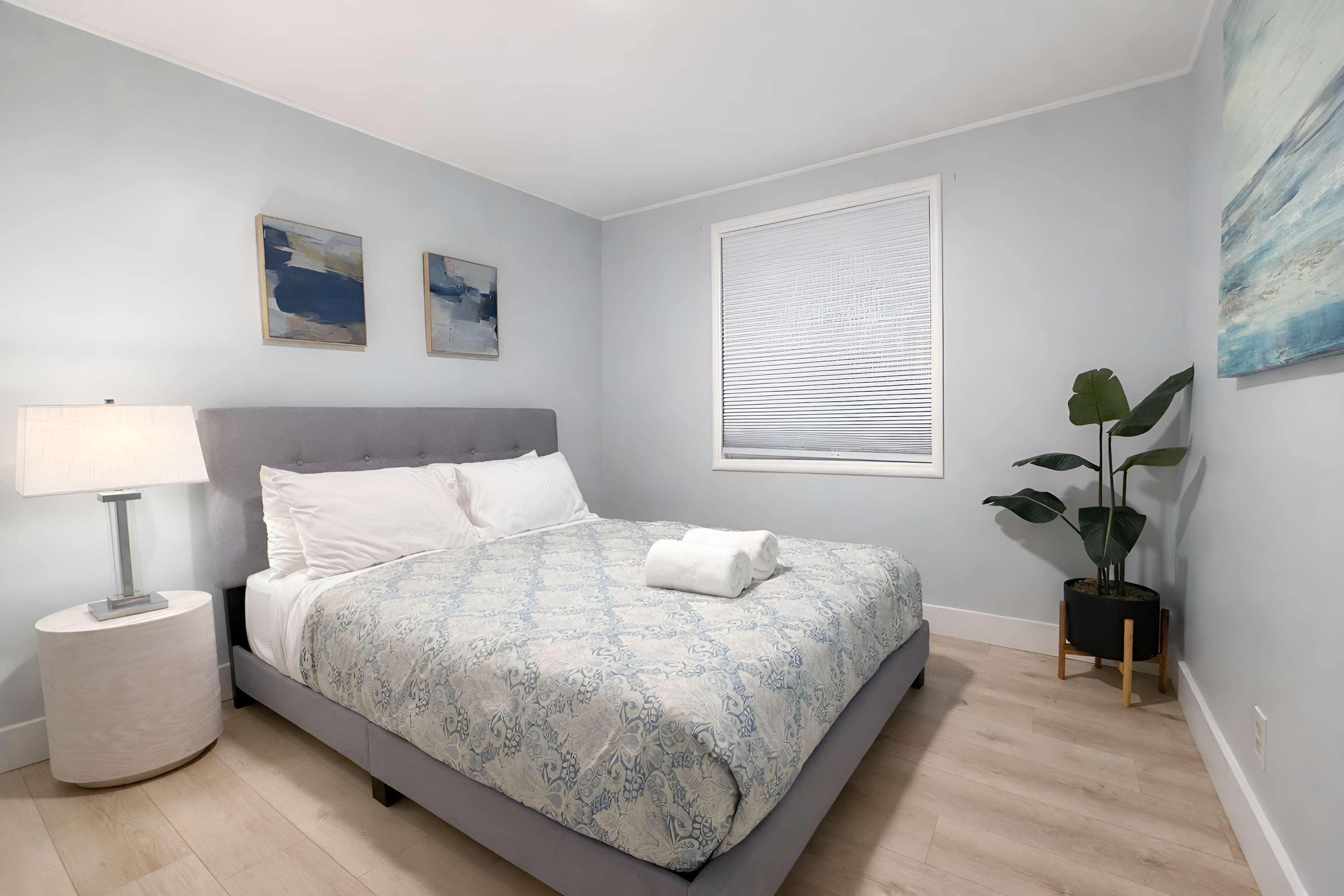 A bedroom with a bed, a nightstand, a lamp, a window with blinds, and a plant.