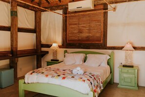 Een kamer met een groen bed, twee nachtkastjes met lampen, een kleine koelkast en een raam met houten luiken.