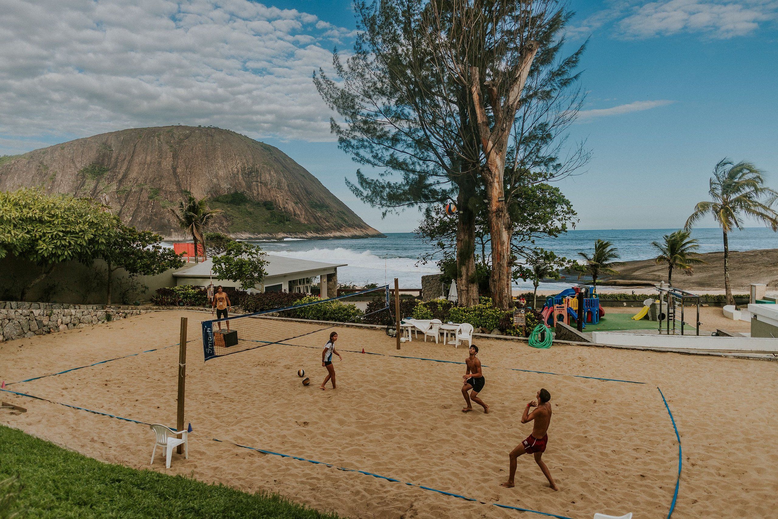 Una cancha de voleibol de playa con una red, rodeada de arena, palmeras y una montaña al fondo.