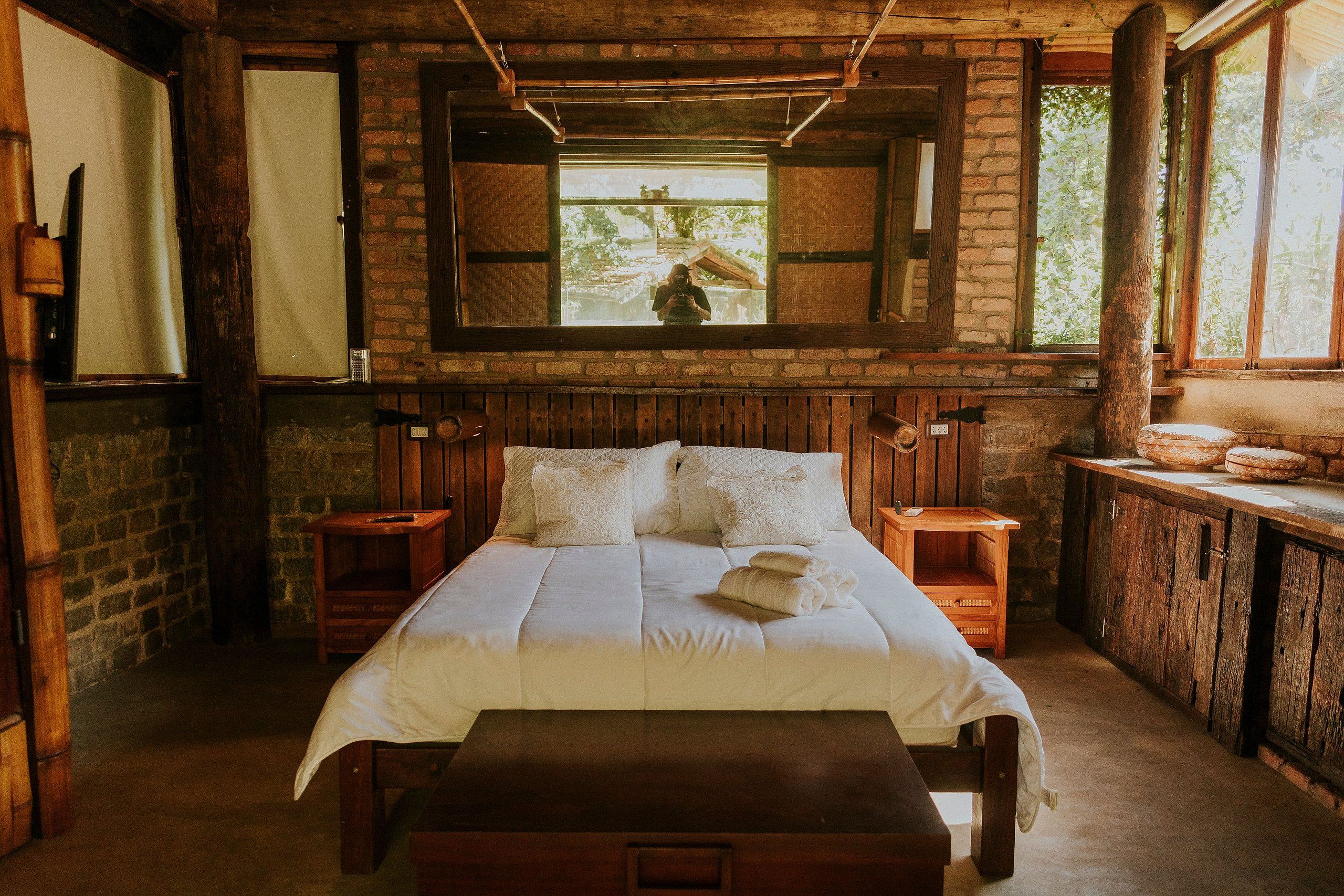 Una habitación rústica de madera con una cama grande, mesitas de noche de madera, un banco y una ventana con vistas a la vegetación.