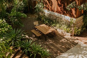 Una terraza de madera con una mesa y bancos, rodeada de plantas diversas y un muro de piedra.