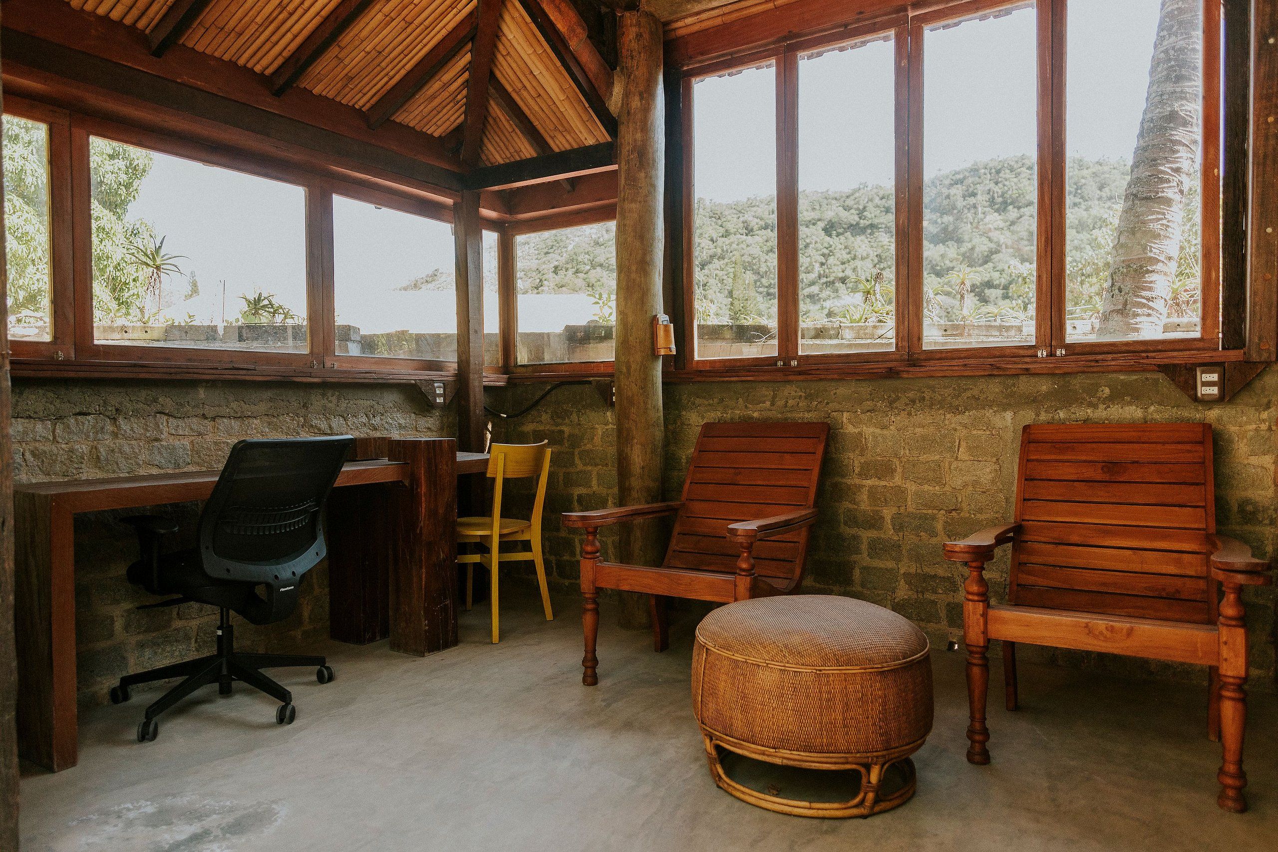 Une pièce comprenant un bureau en bois, deux chaises en bois, un pouf en osier et de grandes fenêtres donnant sur un paysage montagneux.