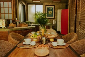 A breakfast setup with wicker chairs, a wooden table, and a variety of food and drink items. - Ypê Greenhouse (Niteroi)