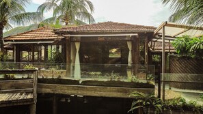 A traditional wooden structure with a tiled roof, surrounded by lush greenery and palm trees. - Ypê Greenhouse (Niteroi)