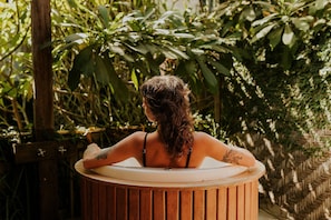 Iemand ontspant in een houten jacuzzi, omringd door weelderige begroeiing.