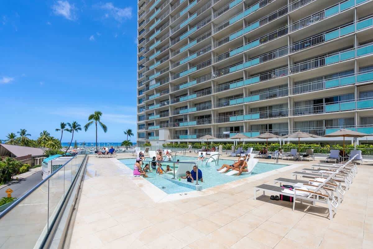 A hotel pool area with a large pool, lounge chairs, and a high-rise building in the background.