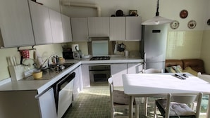 Een kleine keuken met witte kasten, een gasfornuis, een inbouwoven en een koelkast. In de aangrenzende woonkamer staat een eettafel met stoelen en een kleine bank.