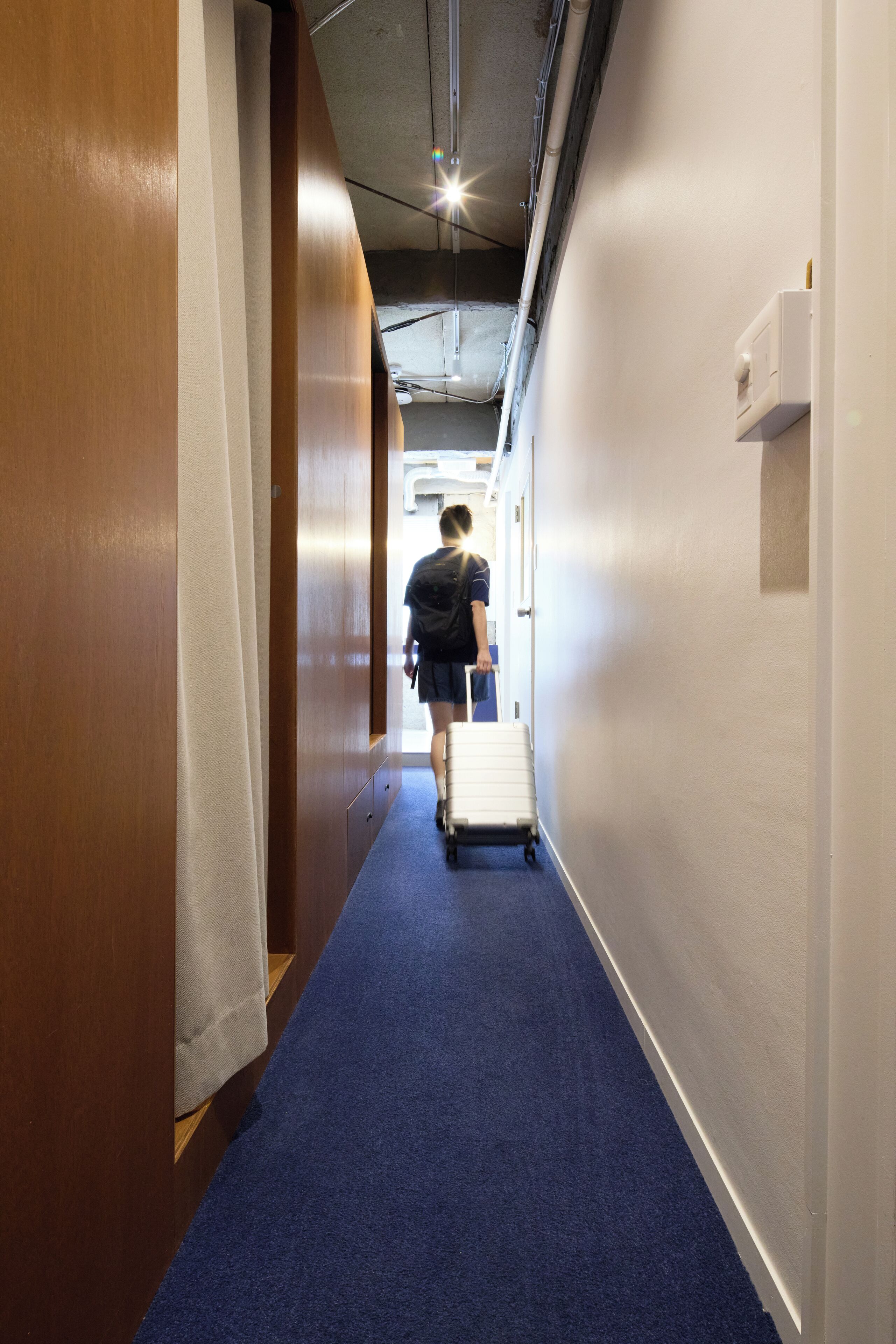 A narrow hallway with a blue carpet, wooden walls, and a person pulling a suitcase.