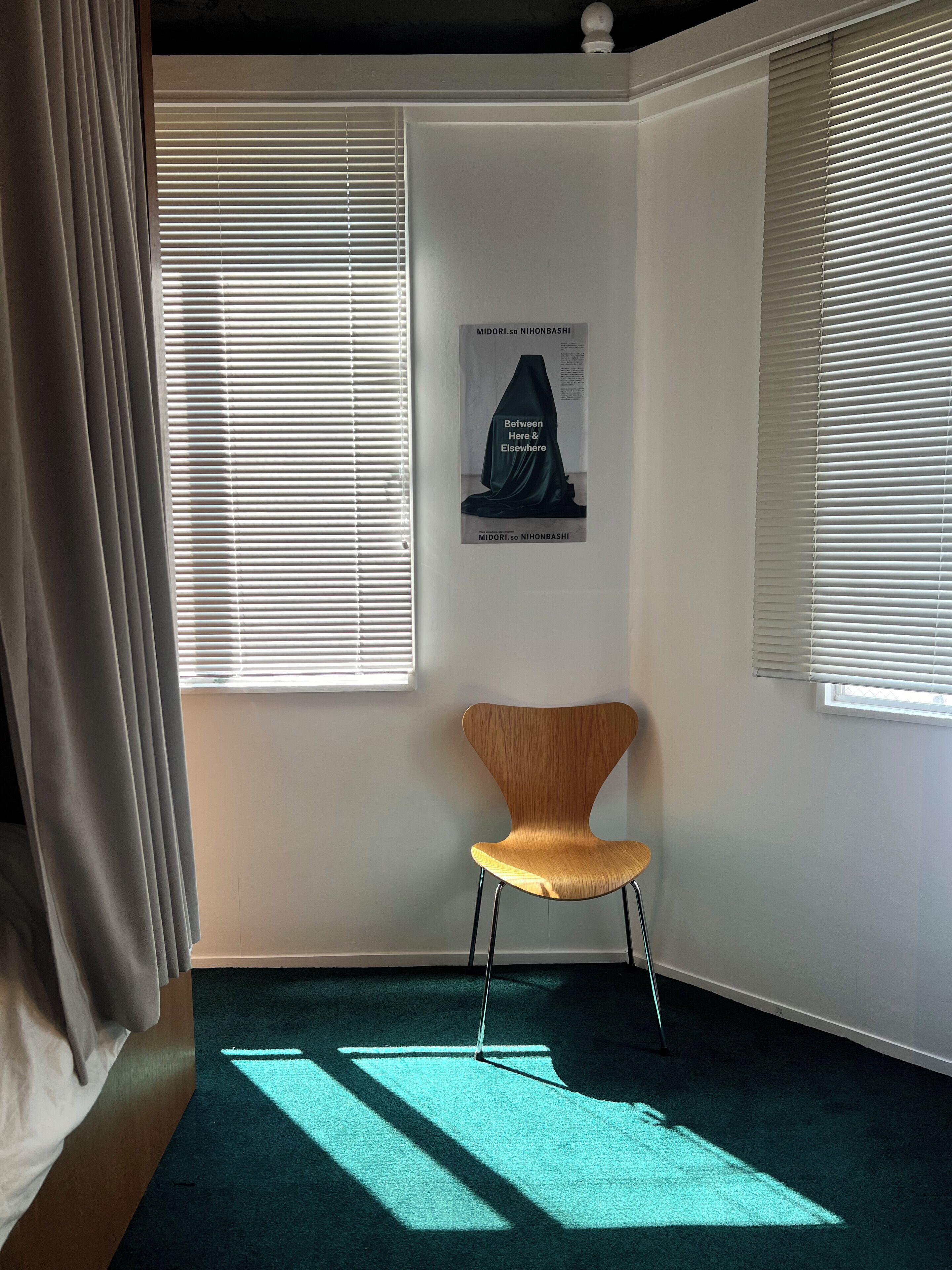 Een kamer met een enkele stoel, twee ramen met jaloezieën, een poster aan de muur en een gedeeltelijk geopend gordijn.