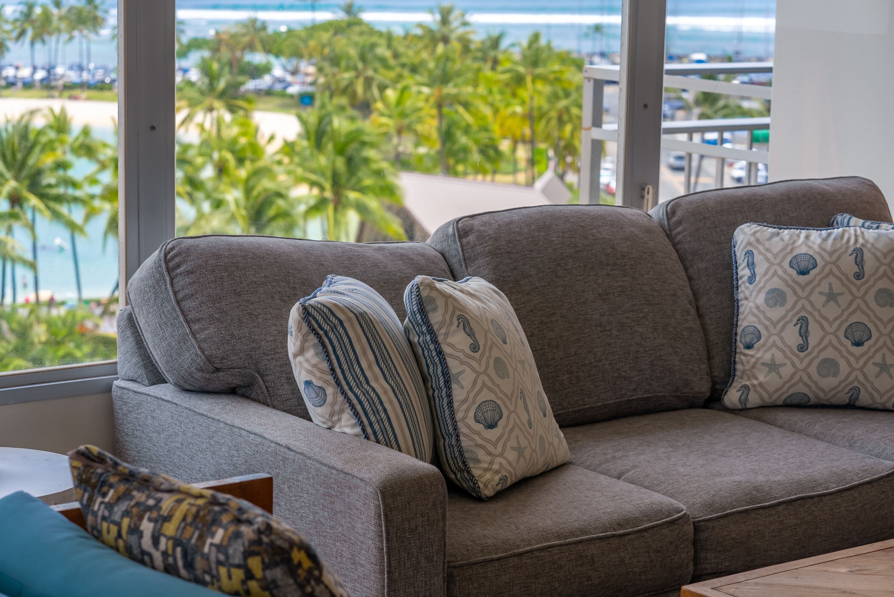 A grey sofa with patterned cushions, a view of a beach and palm trees through large windows.